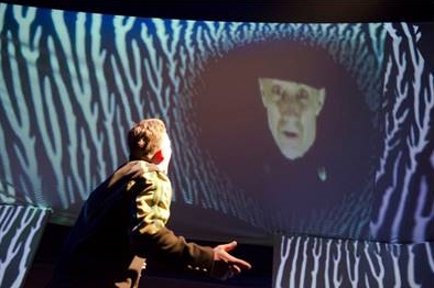 Bild aus einem Theaterstück mit einem Mann und einer Videoprojektion.
