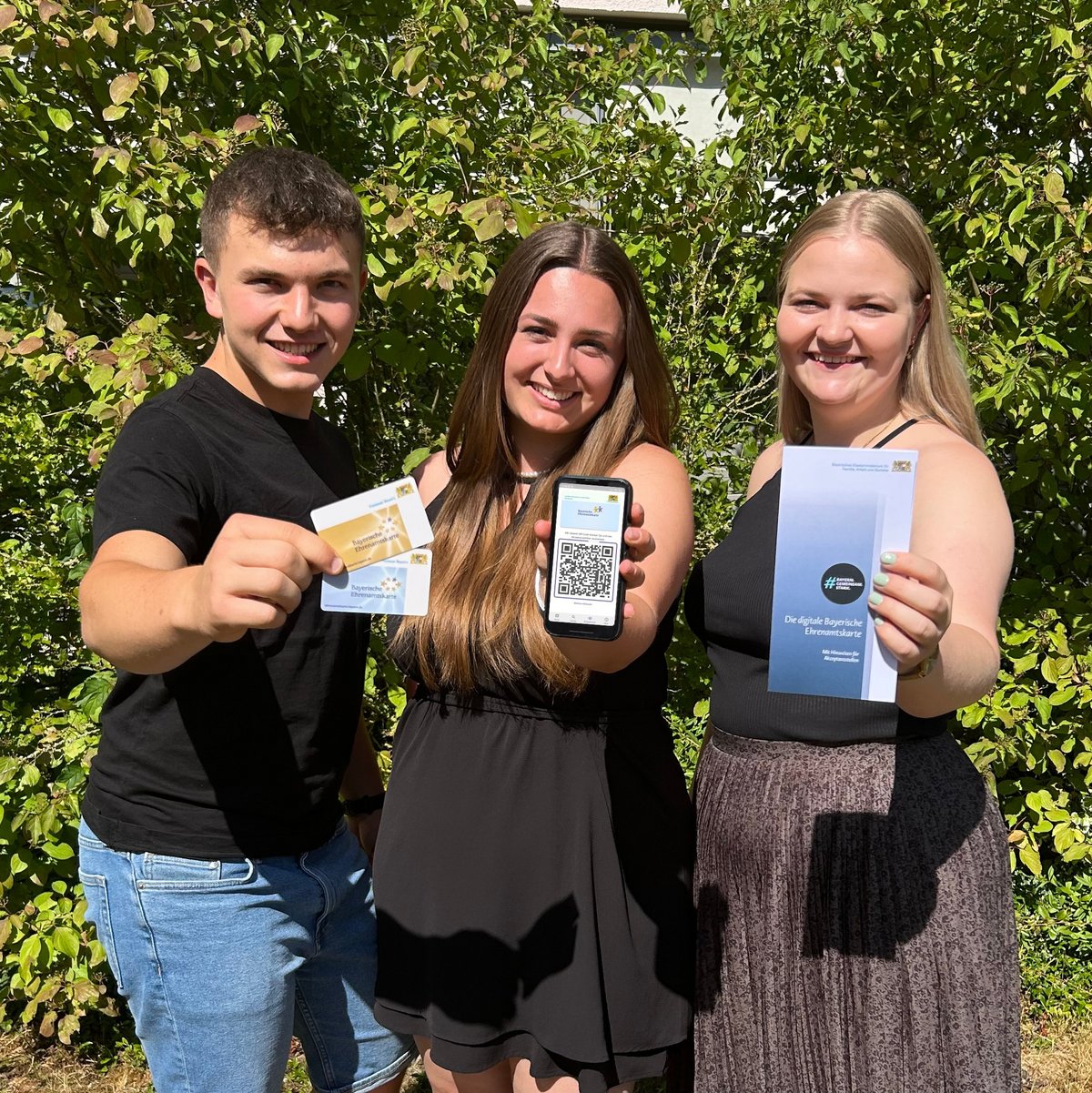 Drei junge Personen halten blaue und goldene Ehrenamtskarte, ein Smartphone und einen Flyer zur Ehrenamtskarte  in die Kamera